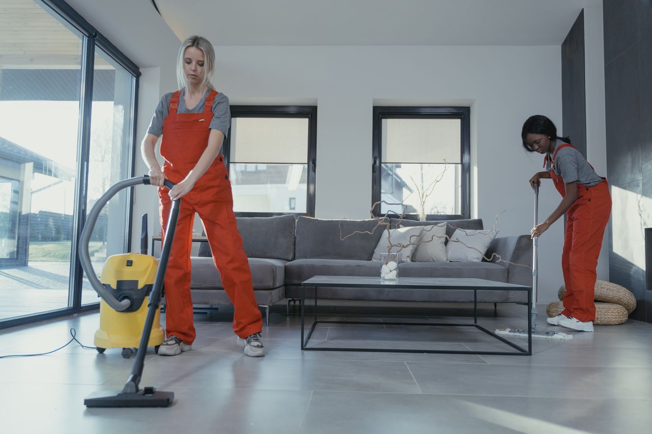 team-03 Two women in red overalls vacuuming and sweeping a modern living room.