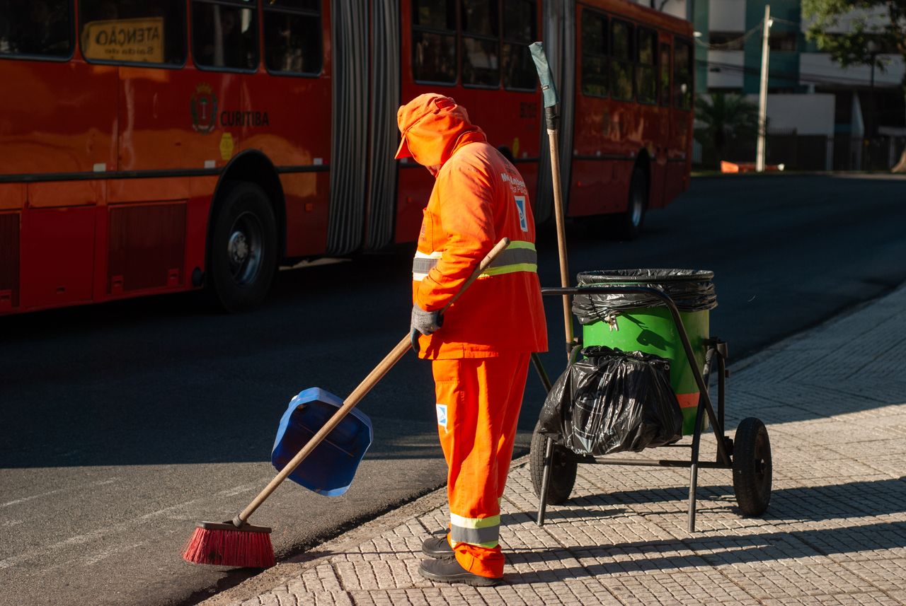 team-02 Street cleaner in bright orange uniform sweeping sidewalk near public bus in city setting.